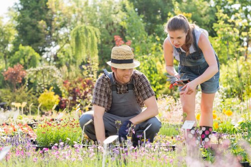 A gardener and a young girl are engaged in gardening activities within a well-maintained garden, with lush green trees and colorful flowering plants in the background. The gardener, wearing a straw hat, a checked shirt, and gardening gloves, is kneeling on the grass near a flower bed, tending to the plants. The girl, dressed in denim overalls and a sleeveless top, is standing beside the gardener, holding garden tools and assisting with the planting or pruning process. The garden features a neatly edged flower bed with an array of vibrant flowers and a manicured lawn with evenly trimmed grass. In the background, there are dense trees including a weeping willow and other leafy shrubs, creating a lush, natural setting typical for outdoor gardening in the Yeading area. The scene appears to be set on a sunny day, with natural light casting soft shadows across the garden. This outdoor space reflects professional gardening and landscaping practices, suitable for garden maintenance and hedge trimming services offered by Hedgetrimmingyeading.co.uk as part of comprehensive outdoor care in the local area.