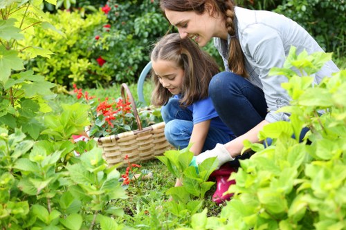 A garden scene in Yeading showing a young woman and a girl engaging in outdoor gardening activities. The foreground features lush green foliage, including a variety of dense, leafy plants and small flowering bushes with red blossoms. Behind them, a well-maintained grass lawn is visible, bordered by a neatly trimmed hedge that runs along the perimeter of the garden. The hedge, likely a common species used in local landscaping, has a uniform height and dense foliage, indicating recent trimming by professionals such as those from Hedge Trimming Yeading. To the right, a paved patio area with textured grey stones integrates into the garden layout, providing a clear outdoor seating or activity space. The overall setting appears bright, under natural daylight with gentle shadows, suggesting fair weather conditions. The scene captures a peaceful, well-kept outdoor environment typical of residential gardens in the Yeading area, highlighting elements that would benefit from regular gardening and hedge-trimming services to maintain its neat and inviting appearance.